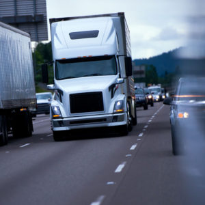 Modern big rig semi truck in the middle of a busy traffic at night with the headlights on highway and collon cars moving back and side of the semi truck with a refrigerator trailer unit.